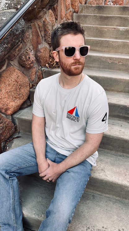 A man with a beard and sunglasses is sitting on outdoor stone steps, wearing a light silver t-shirt with a red and blue Divine Mercy logo on the left chest. Catholic Clothing.