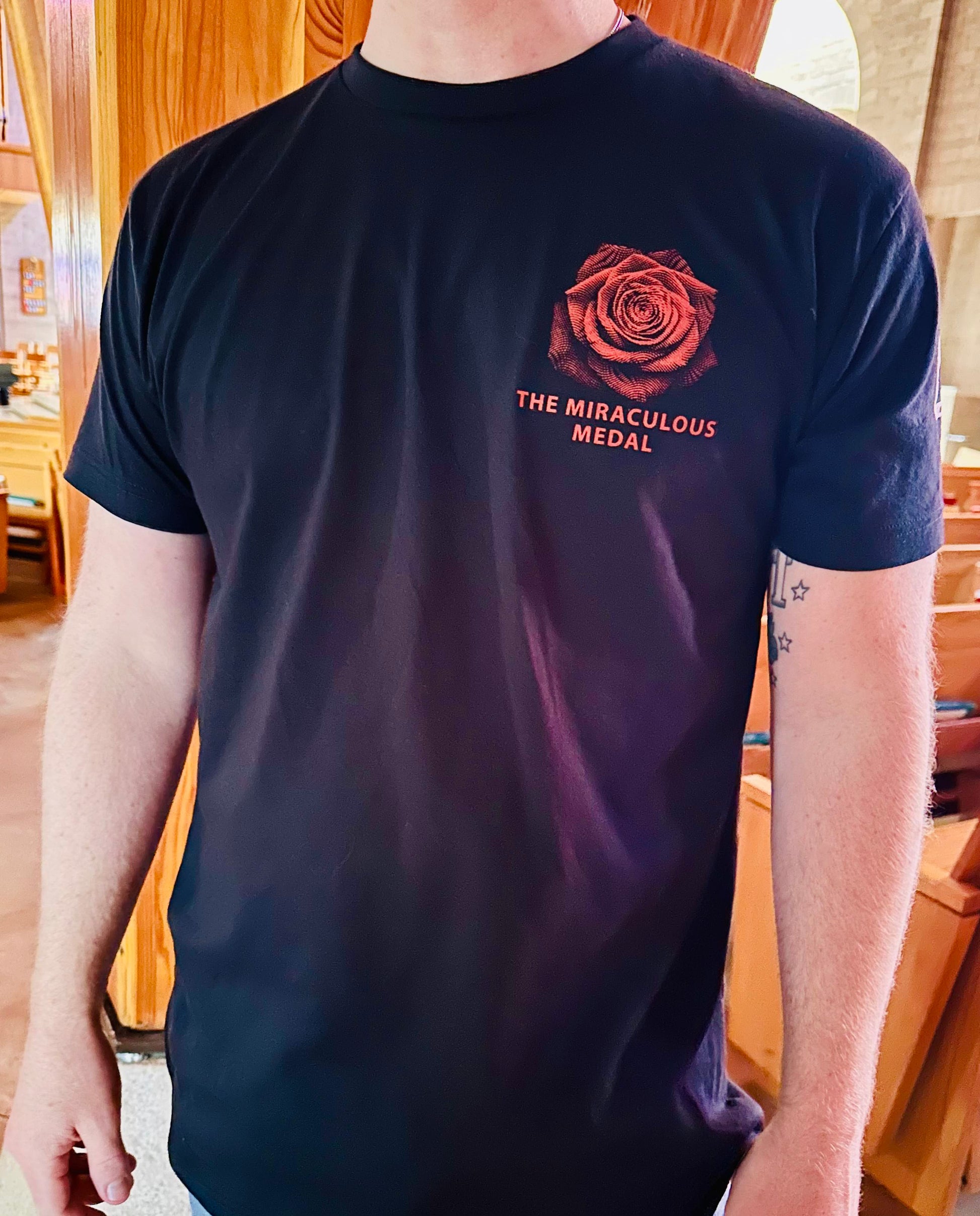 A man wearing a black crewneck T-shirt with a red rose graphic and the text 'THE MIRACULOUS MEDAL' printed on the left chest, set against a warm indoor Catholic Church. Catholic Clothing.