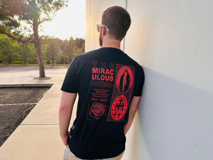 A man wearing a black T-shirt with a large red graphic of the Miraculous Medal printed on the back, photographed from behind as he leans against a white wall outdoors. Catholic Clothing.