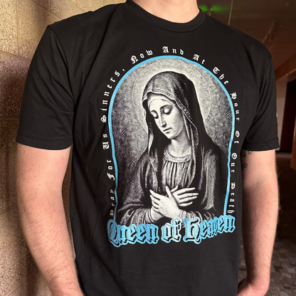 A person wearing a black short-sleeve graphic T-shirt featuring a large front print of the Virgin Mary in grayscale, surrounded by a blue oval border and the words 'Queen of Heaven' in blue gothic lettering.