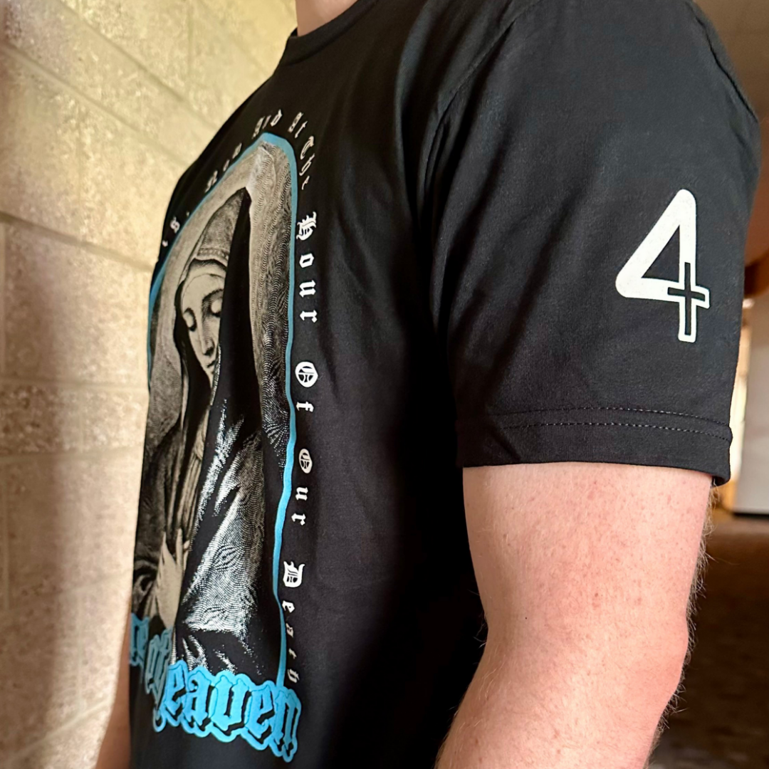 A close-up of a person wearing a black graphic t-shirt. The shirt features a large grayscale illustration of the Virgin Mary with bright blue and white gothic text, and a white number 4 with a cross on the right sleeve. Catholic Clothing.