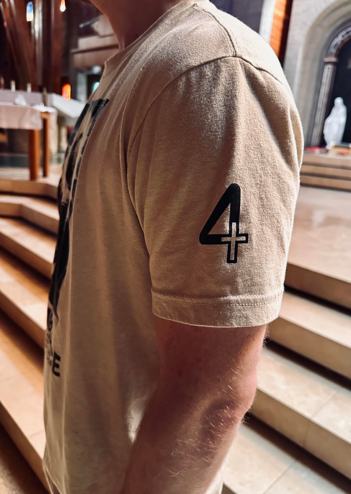 A close-up side view of a person wearing a beige T-shirt with a black graphic print of the number 4 featuring a cross on the sleeve, set against a blurred background of stone steps in a Catholic Church. Catholic Clothing.