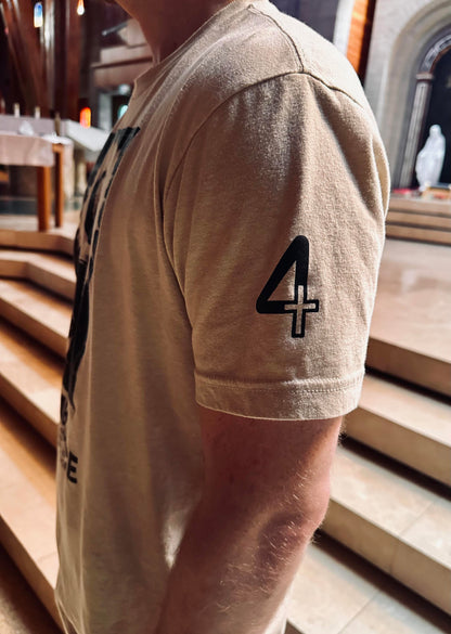 A close-up side view of a person wearing a beige T-shirt with a black graphic print of the number 4 featuring a cross on the sleeve, set against a blurred background of stone steps in a Catholic Church. Catholic Clothing.