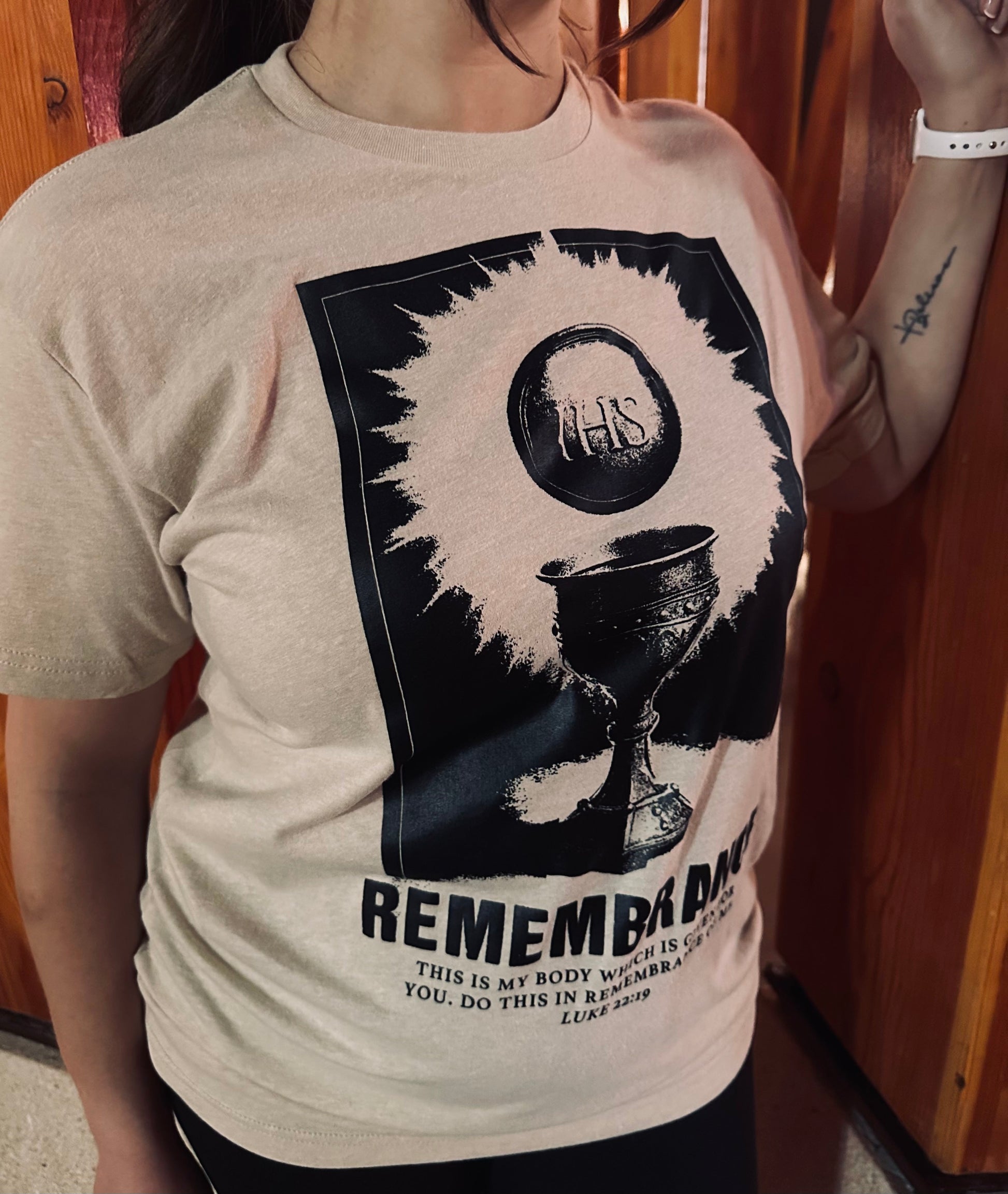 A woman wearing a beige graphic t-shirt with a black print of The Catholic Eucharist with the words Remembrance, and the blble verse, Luke 22: 19. Catholic Clothing.