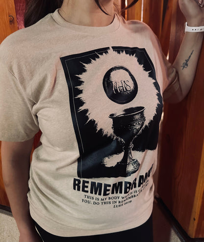 A woman wearing a beige graphic t-shirt with a black print of The Catholic Eucharist with the words Remembrance, and the blble verse, Luke 22: 19. Catholic Clothing.