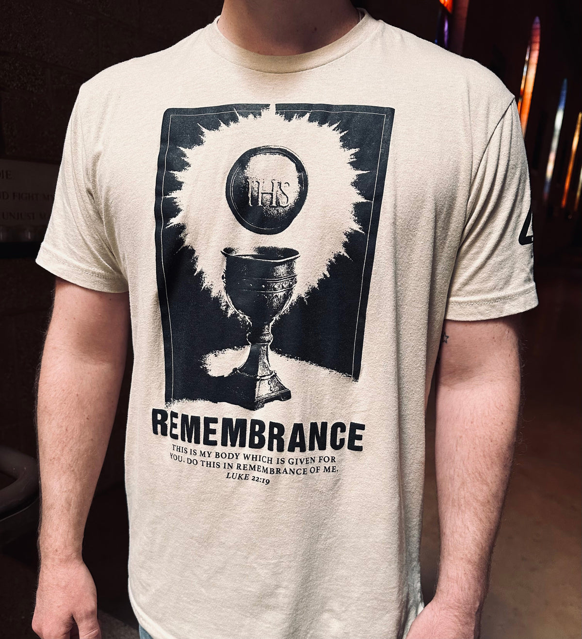 A man wearing a light beige t-shirt with a large black graphic print of the Eucharist (chalice and IHS monogram) with the word 'REMEMBRANCE' printed below.