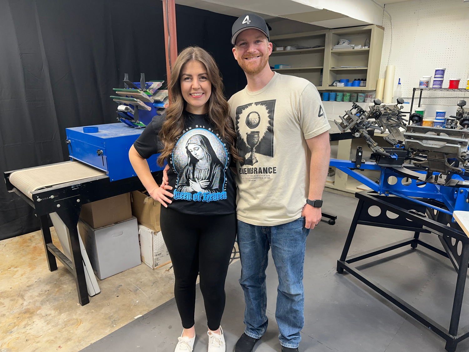Picture of Threads 4 Catholics owners, Nate and Stephanie wearing the remembrance eucharist t-shirt and the queen of heaven t-shirt. Photo taken in the threads 4 catholics screen printing shop.